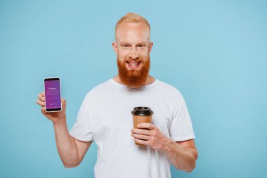 Kyiv, Ukrayna - 27 Ağustos 2019: Instagram uygulamalı akıllı telefonu göstermek için elinde kahveyle gülümseyen sakallı adam, mavi üzerine izole edilmiş.