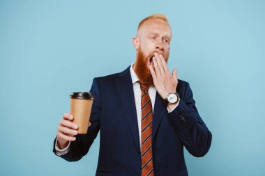 tired bearded businessman in suit yawning and holding coffee to go, isolated on blue clipart