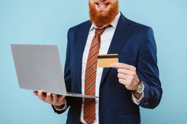 Takım elbiseli gülümseyen sakallı işadamının internetten kredi kartı ve laptopuyla alışveriş yapması, mavi renkte izole edilmiş.