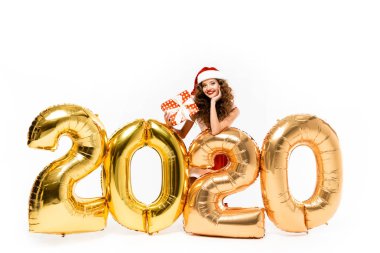 Noel Baba kostümlü mutlu bir kız elinde hediye kutusuyla 2020 yılındaki altın balonlarla poz veriyor.