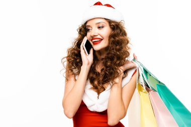 Noel Baba kostümü giymiş gülümseyen kıvırcık kız alışveriş torbaları tutuyor ve akıllı telefondan konuşuyor, beyaz tenli.