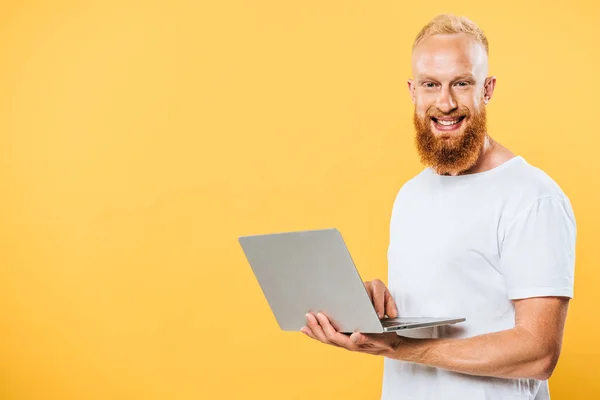 smiling bearded man using laptop, isolated on yellow