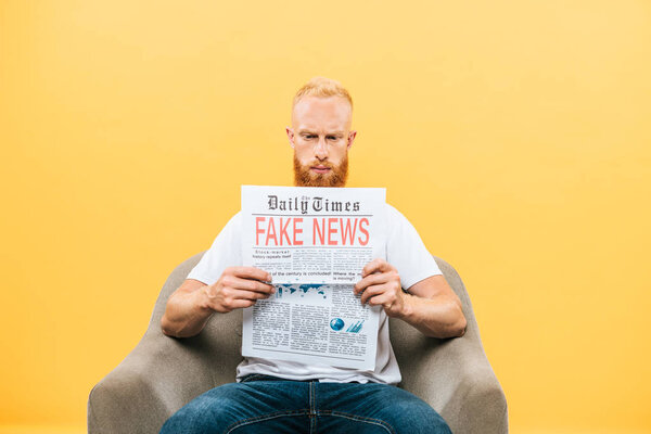 serious man reading newspaper with fake news while sitting on armchair, isolated on yellow
