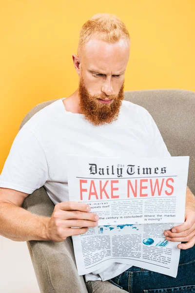 serious man reading newspaper with fake news while sitting on armchair, isolated on yellow