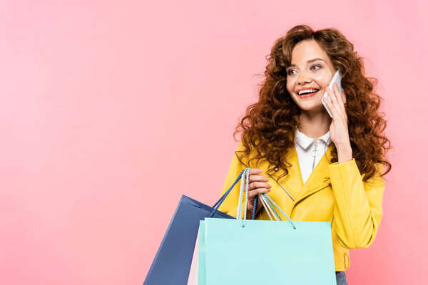beautiful curly girl holding shopping bags while talking on smartphone, isolated on pink