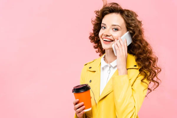 happy curly girl with coffee to go talking on smartphone, isolated on pink