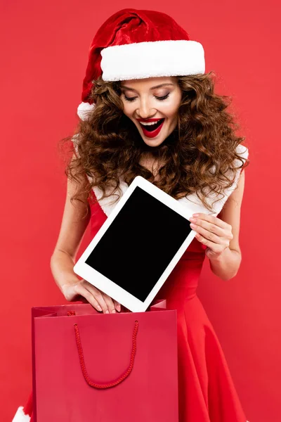 兴奋的卷曲女孩 身穿圣塔服装 手持装有数码平板电脑的购物袋 与红色隔离 — 图库照片