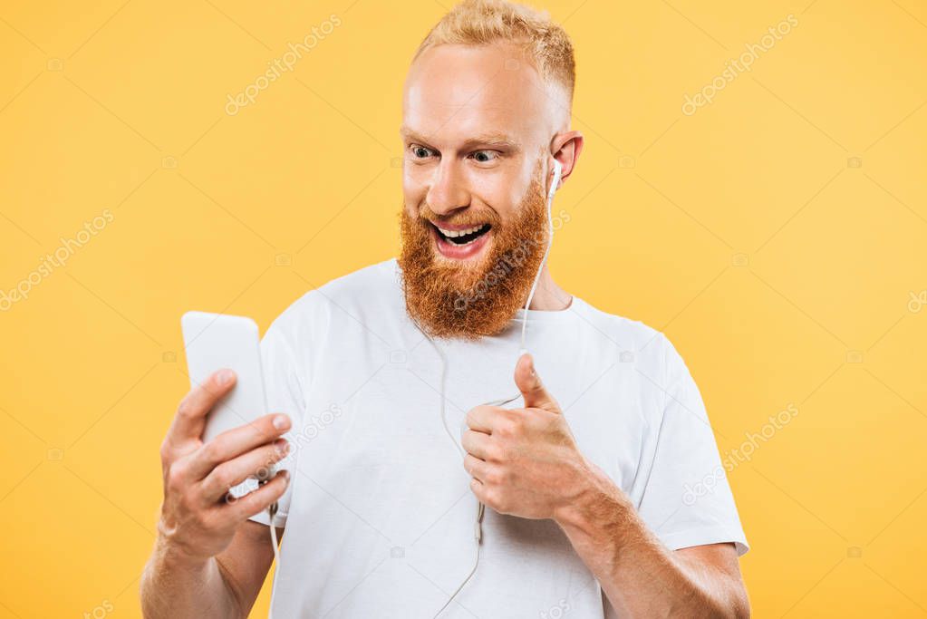 cheerful bearded man with earphones taking selfie on smartphone and showing ok sign, isolated on yellow