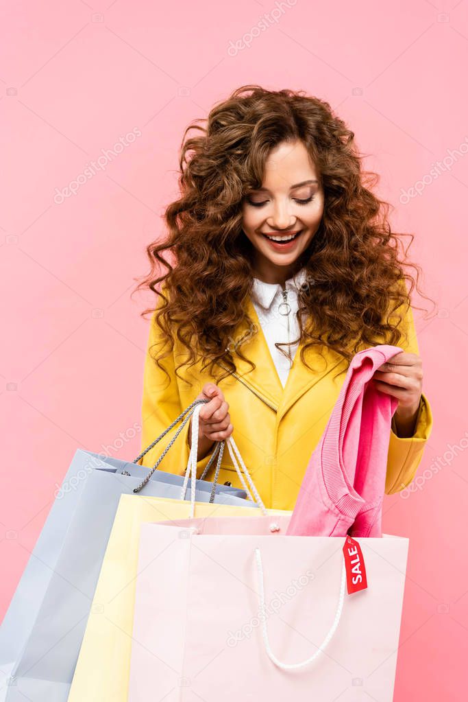 beautiful curly girl taking clothes from shopping bags with sale tag, isolated on pink