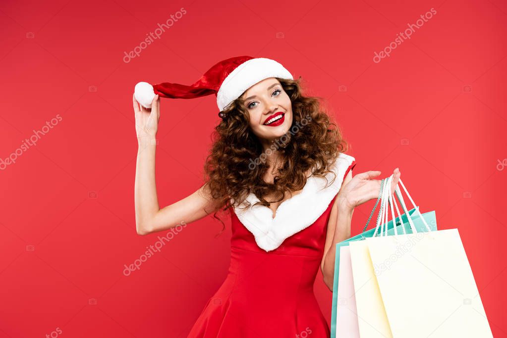 happy curly girl in santa costume and hat posing with shopping bags, isolated on red