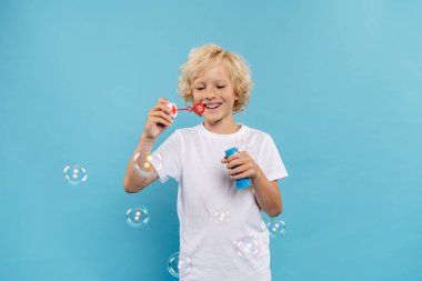 Beyaz tişörtlü gülümseyen çocuk mavi arka planda sabun köpüğü üflüyor. 
