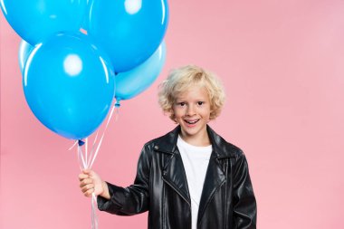 Gülümseyen çocuk elinde balonlarla pembe kameraya bakıyor.
