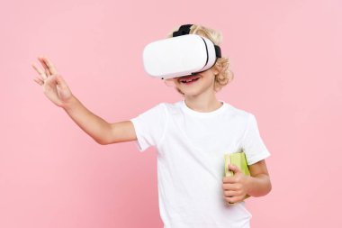 Yüzünde sanal gerçeklik kulaklığı olan gülümseyen bir çocuk pembe üzerine izole edilmiş kitap tutuyor.