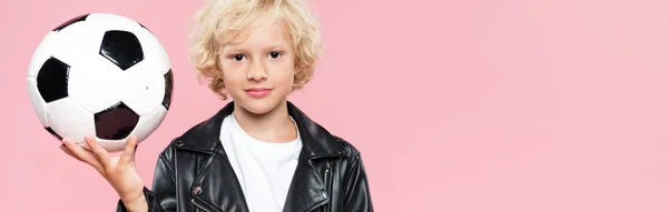 panoramic shot of kid in leather jacket holding football and looking at camera isolated on pink