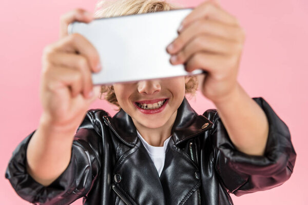 selective focus of smiling kid holding smartphone isolated on pink