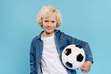 Gülümseyen ve sevimli bir çocuk kameraya bakıyor ve mavi üzerine izole futbol topu tutuyor. 