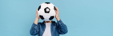 Mavi üzerine izole edilmiş futbolla yüzünü gizleyen çocuğun panoramik görüntüsü. 