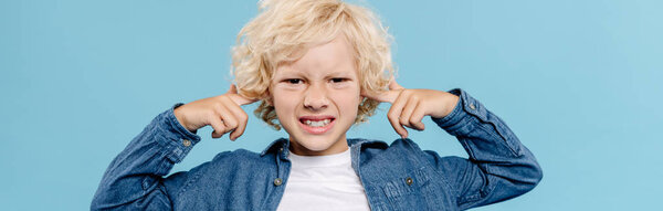 panoramic shot of irritated and cute kid looking at camera and touching ears isolated on blue 