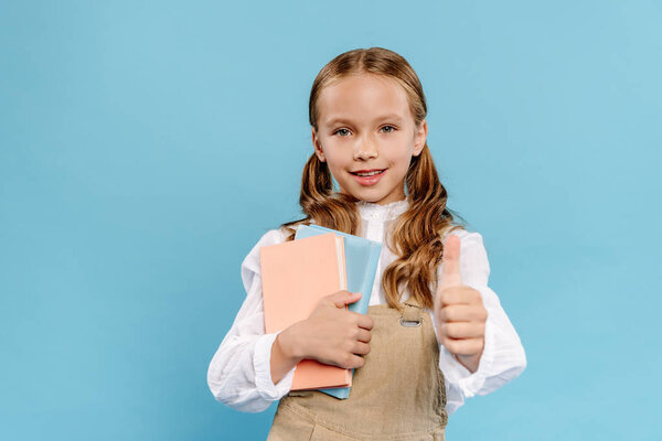 smiling and cute kid showing like gesture and holding books isolated on blue 