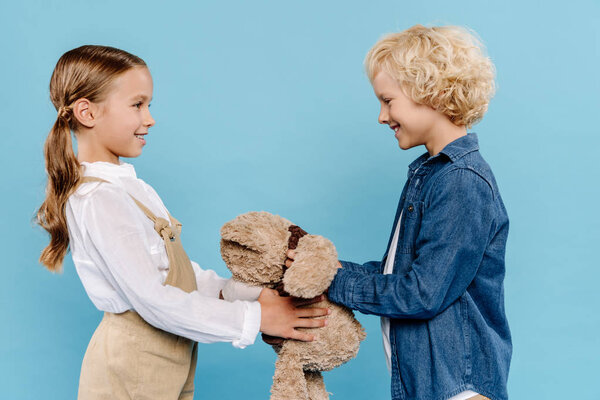 side view of smiling and cute kids holding teddy bear isolated on blue 