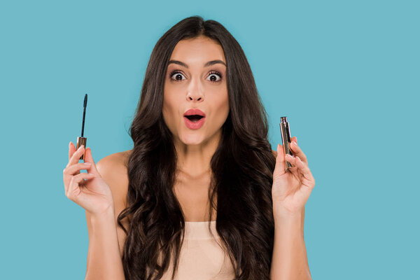 young and surprised woman holding mascara isolated on blue 