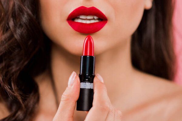 cropped view of woman holding lipstick near red lips isolated on pink 