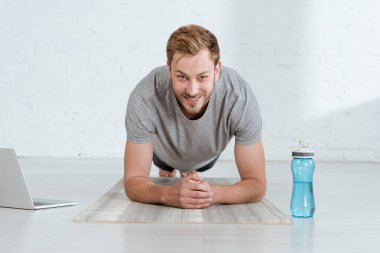 smiling man looking at camera while practicing forearm plank pose near laptop and sports bottle clipart