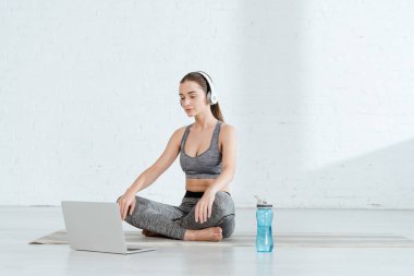 Kulaklıklı genç bir kadın laptopun ve spor şişesinin yanında rahat poz veriyor.