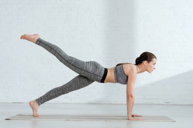 Spor kıyafetli genç bir kadın tek bacaklı tahta duruşuyla yoga yapıyor. 