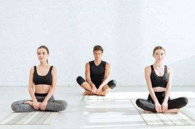 Genç kadınlar ve erkek yarım lotus pozuyla yoga yapıyorlar.