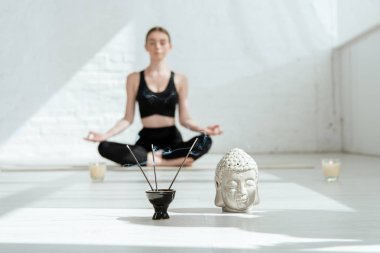 selective focus of young woman sitting in half lotus pose near buddha head sculpture, aromatic sticks and candles clipart