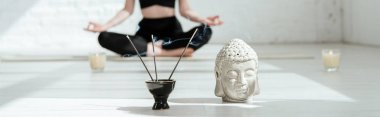 cropped view of woman sitting in half lotus pose near buddha head sculpture, aromatic sticks and candles, panoramic shot clipart