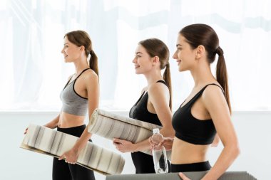 Elinde yoga minderleri tutan üç gülümseyen kadının yan görüntüsü