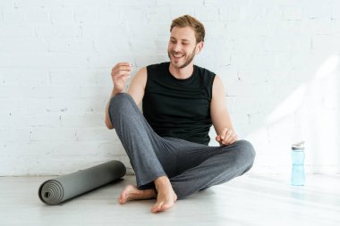 Yoga minderinin, spor şişesinin ve kulaklığın yanında oturan neşeli adam.