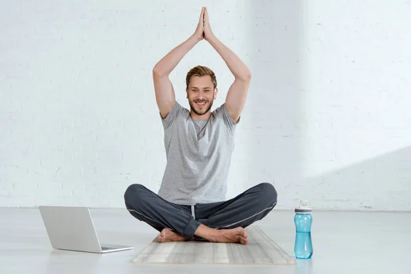 微笑的男人看着相机 一边练习半荷花姿势 一边高举祷告的手靠近笔记本电脑和运动瓶子 — 图库照片