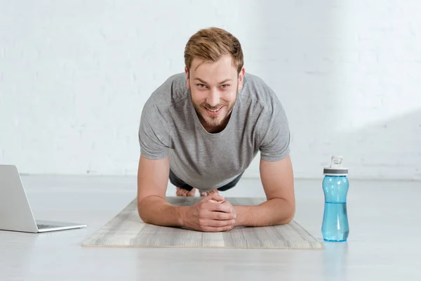 ノートパソコンやスポーツボトルの近くで腕板ポーズを練習しながらカメラを見て笑っている男 — ストック写真