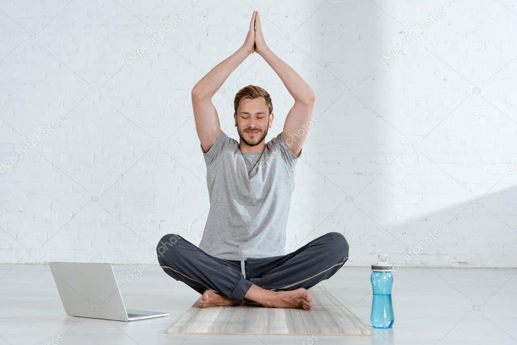young man practicing half lotus pose with raised prayer hands near laptop and sports bottle