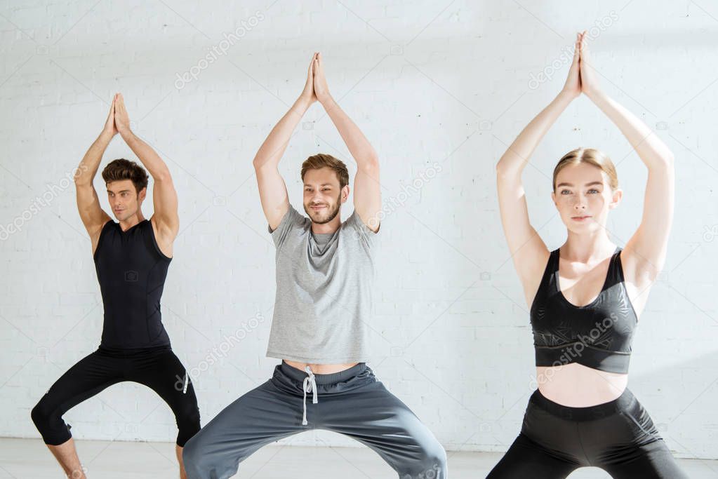 young men and woman practicing yoga in goddess pose with raised prayer hands