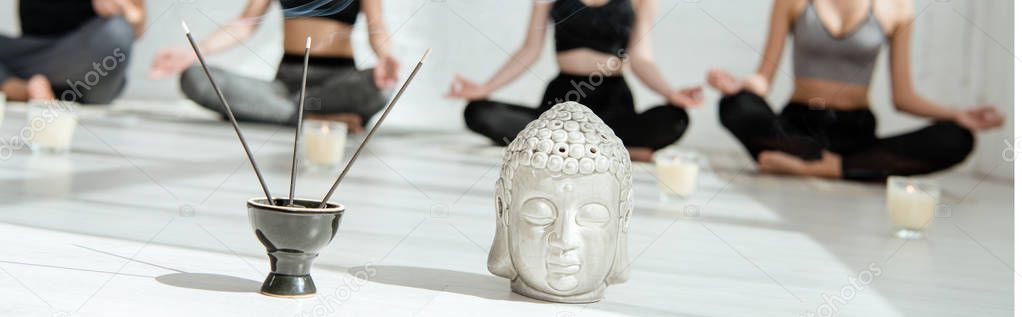 selective focus of decorative buddha head, aromatic sticks and candles, and young people practicing yoga in half lotus pose, panoramic shot