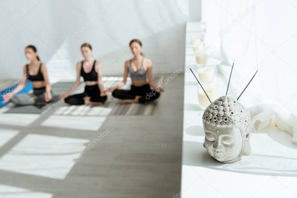 selective focus of Buddha head sculpture with aromatic sticks on windowsill, and three women sitting in half lotus pose