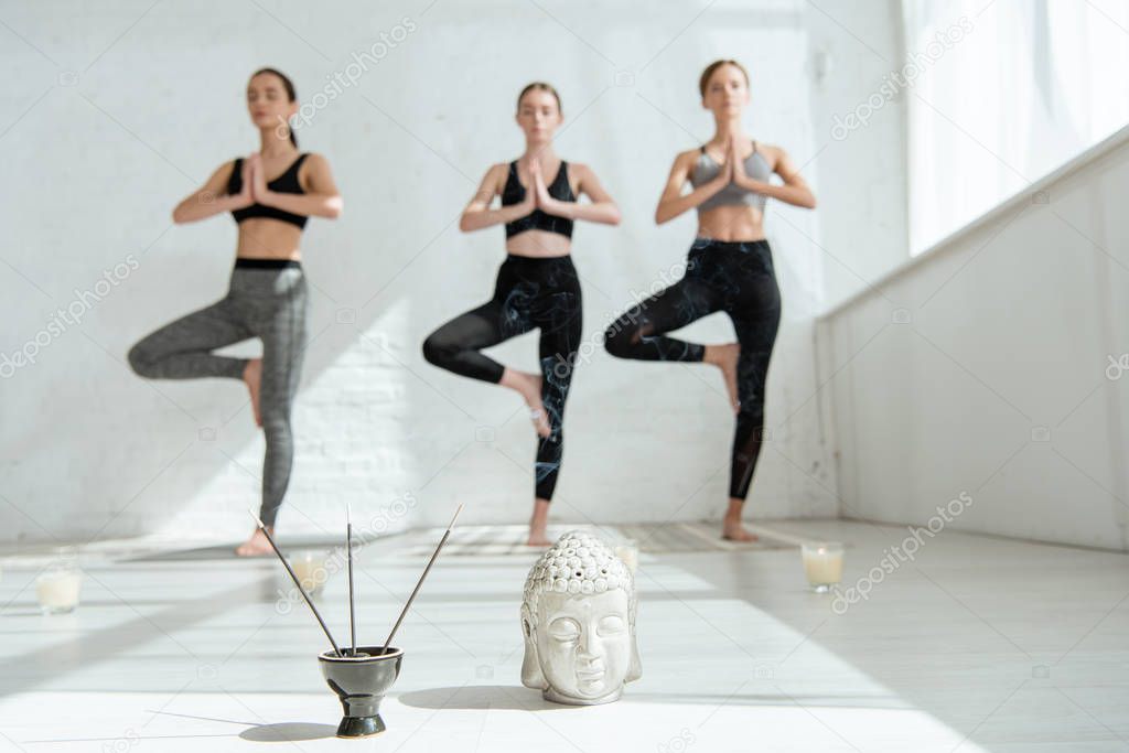selective focus of Buddha head sculpture, incense sticks, and three women standing in tree pose 