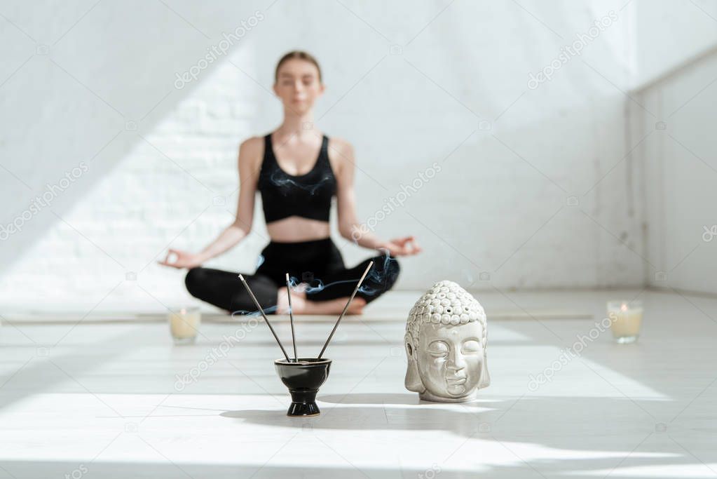 selective focus of young woman sitting in half lotus pose near buddha head sculpture, aromatic sticks and candles