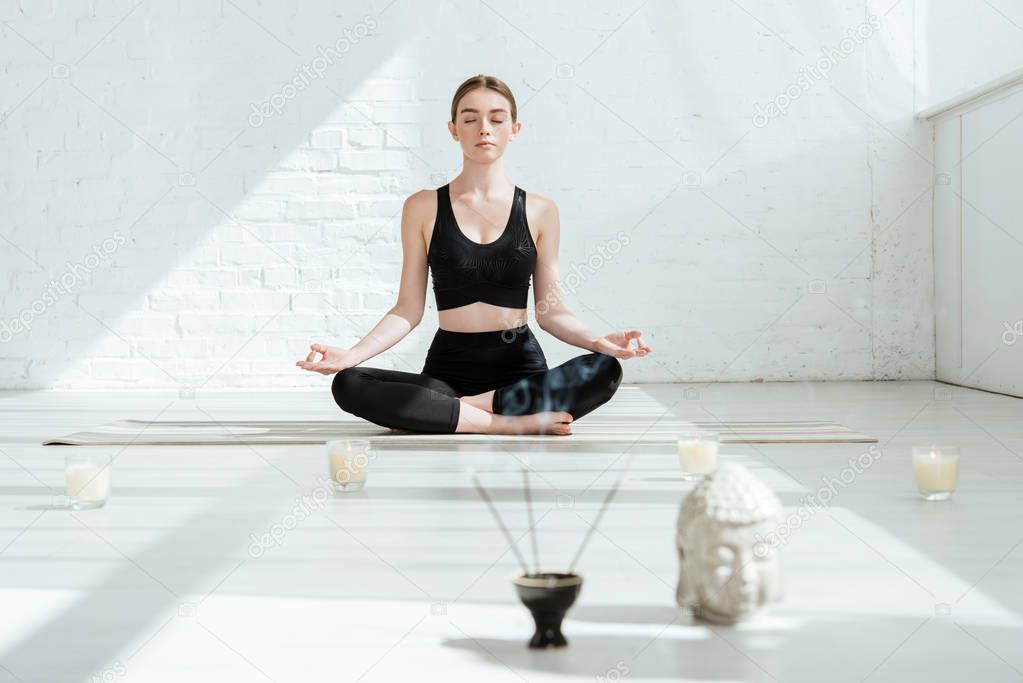 beautiful young woman sitting in half lotus pose near buddha head sculpture, aromatic sticks and candles