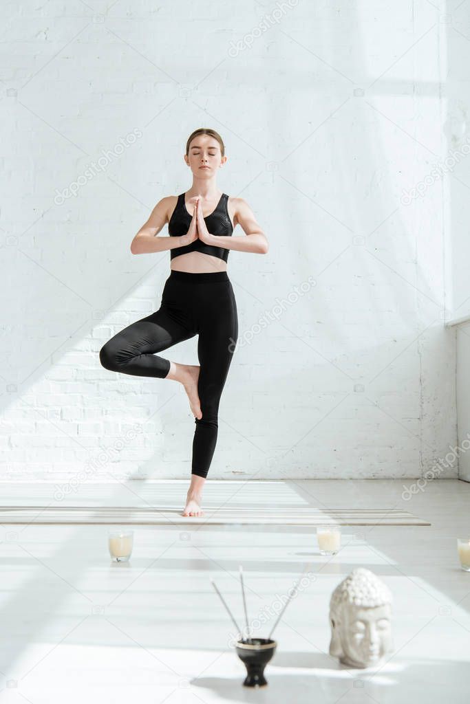 beautiful young woman standing in tree pose near buddha head sculpture, aromatic sticks and candles