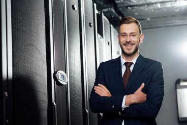 selective focus of cheerful businessman in suit and glasses standing with crossed arms in data center  clipart