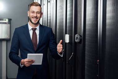 selective focus of cheerful businessman holding digital tablet and showing thumb up in data center  clipart