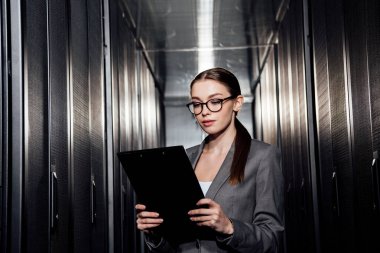 attractive businesswoman in glasses looking at clipboard in data center  clipart