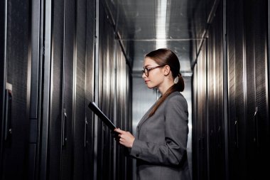 side view of businesswoman in glasses holding clipboard in data center  clipart