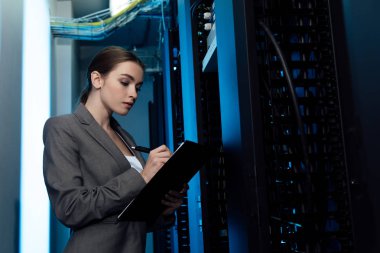 attractive businesswoman writing while holding clipboard in server room  clipart