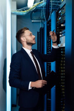profile of handsome businessman in glasses holding pen and clipboard while looking at server rack  clipart